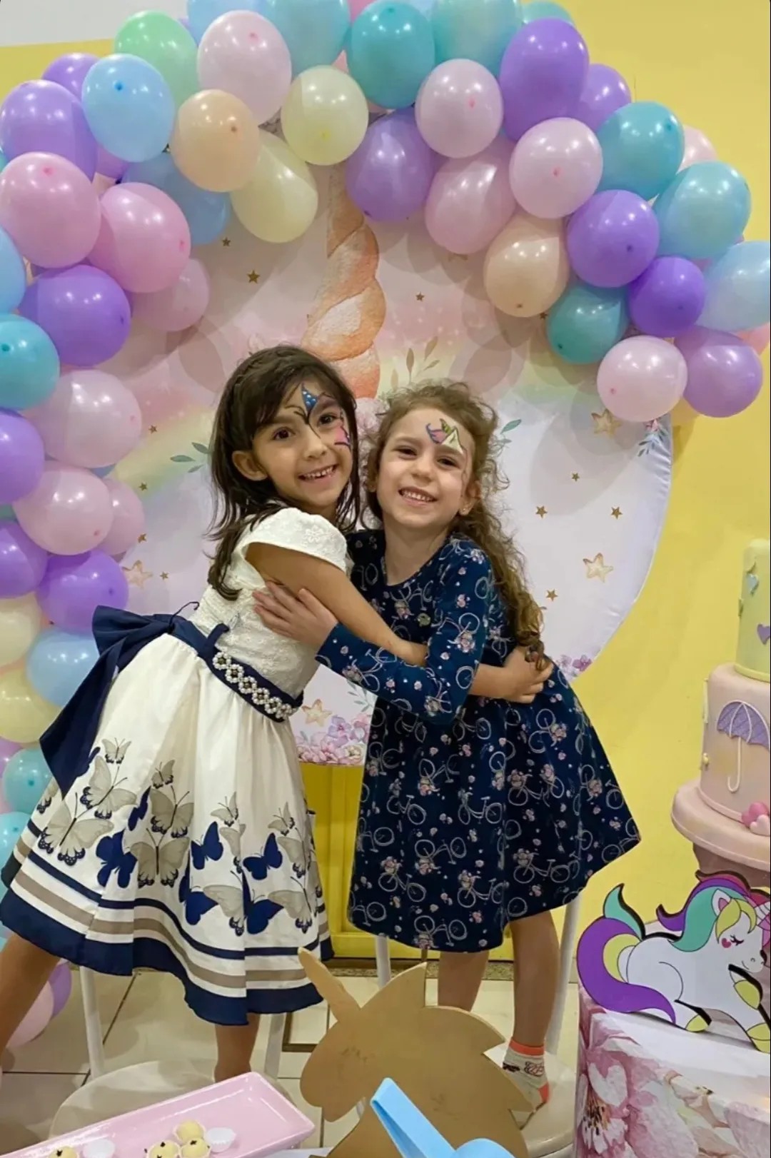 Amigas abraçadas em frente ao painel de unicórnio com balões
